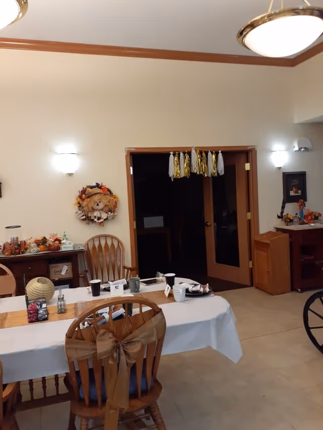 A dining room with a table set for a meal, decorated with a white tablecloth, napkins, cups, and small decorative items. Wooden chairs surround the table, one with a large brown bow tied on the back. The room has beige walls with wooden trim, two wall-mounted lights, a wreath decoration on the wall, and double doors with gold and white tassel decorations hanging above. Additional furniture and autumn-themed decorations are visible in the background.