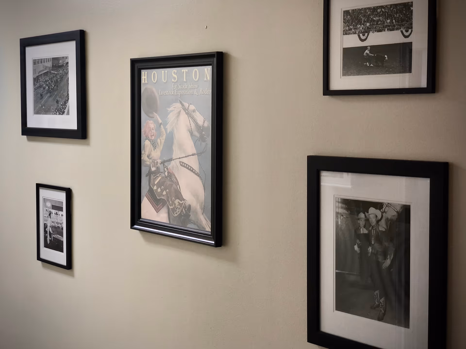 A beige wall with five framed pictures hanging on it. The pictures include black and white photographs and a colored poster featuring a person riding a white horse with the text 'HOUSTON Fat Stock Show Livestock Exposition & Rodeo'.