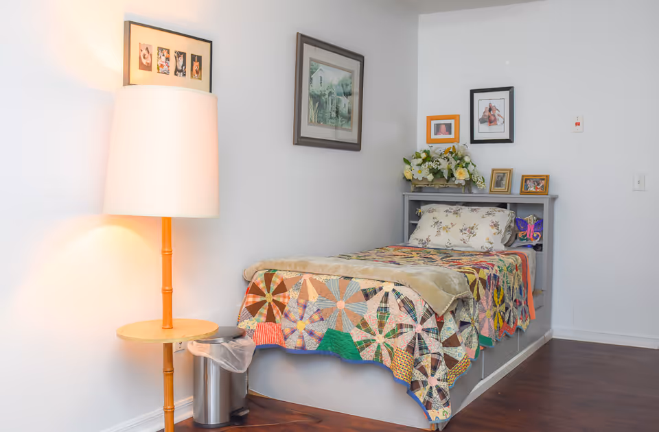 A small bedroom with a single bed covered by a colorful patchwork quilt and a floral pillow. Above the bed are several framed pictures and a floral arrangement on a built-in headboard shelf. To the left of the bed is a wooden floor lamp with a round table attached, next to a small trash can. The room has white walls and dark wooden flooring.