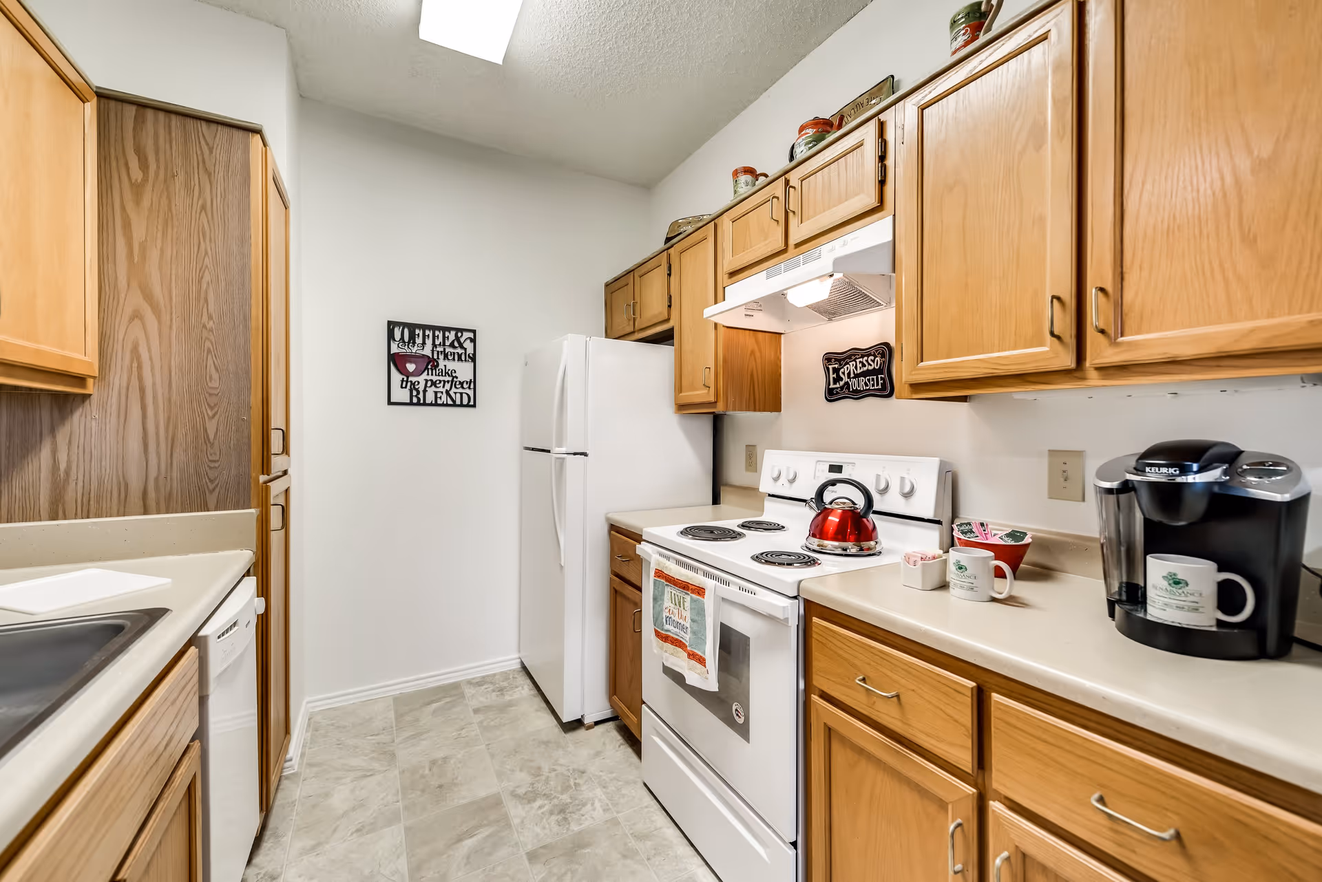 A compact kitchen with wooden cabinets, a white refrigerator, an electric stove with a red kettle on it, a Keurig coffee maker with mugs on the countertop, and decorative coffee-themed signs on the walls.