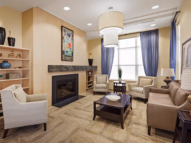 A bright living room with a fireplace, sofa and armchairs arranged around a coffee table in front of a large window with blue curtains.