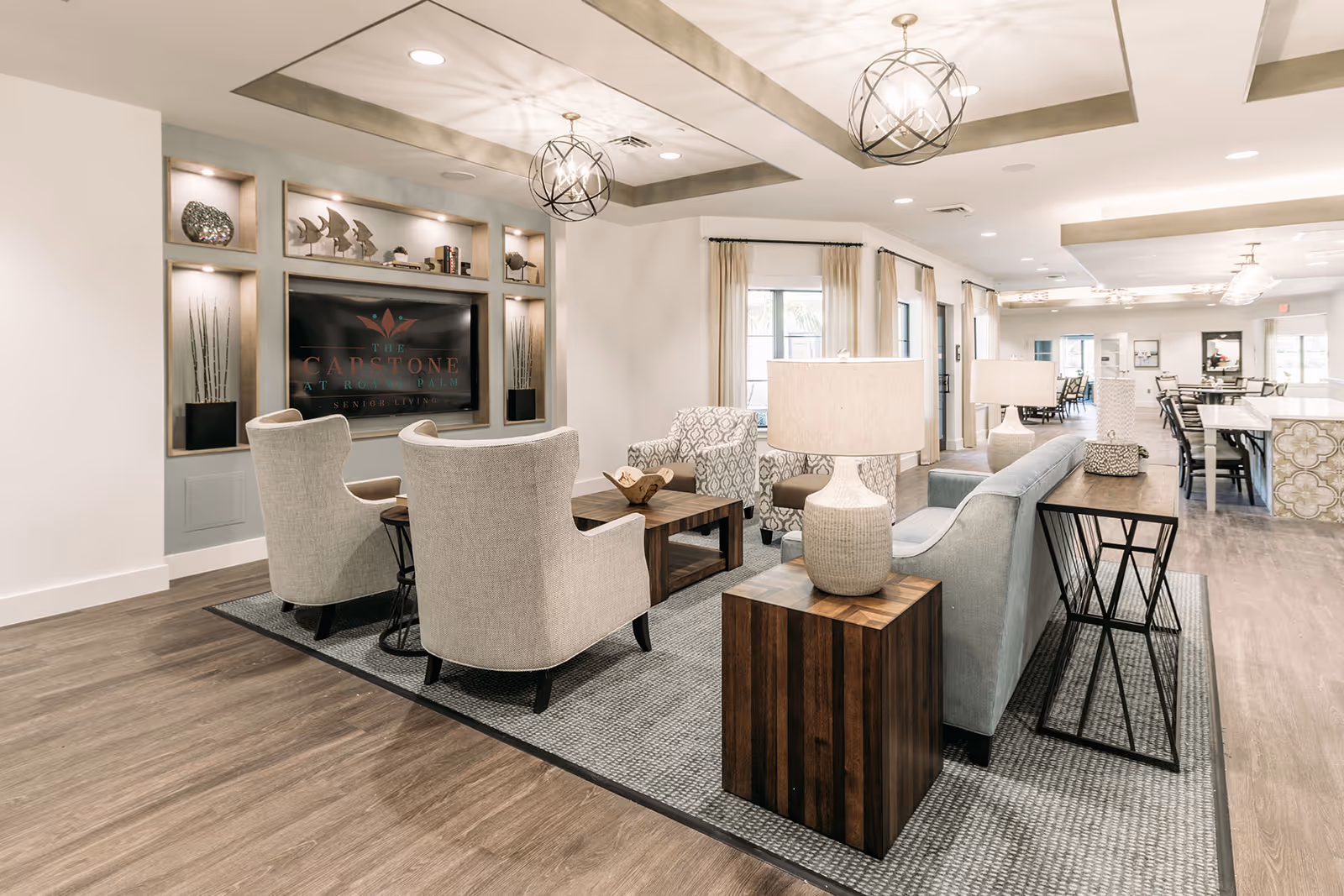 Bright modern communal lounge with sofas, armchairs, lamps, a TV display and dining area in the background.