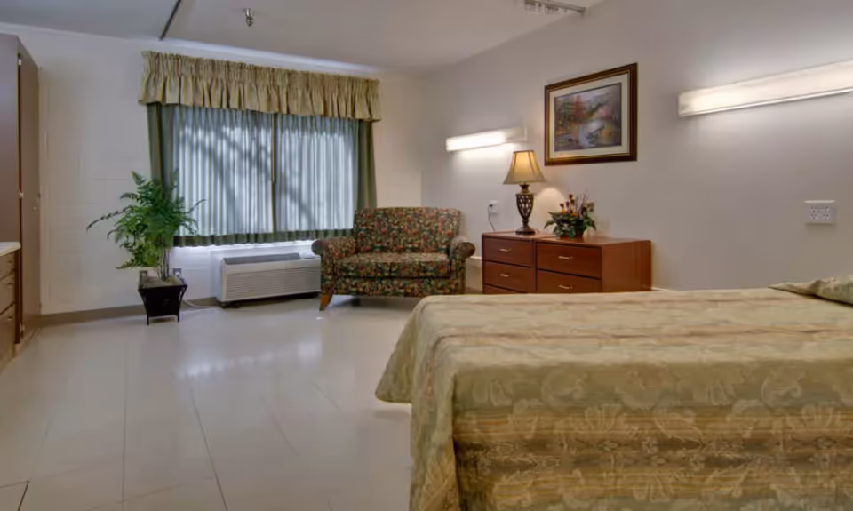 Simple furnished bedroom with a bed in the foreground, floral armchair, dresser with lamp, and a curtained window.