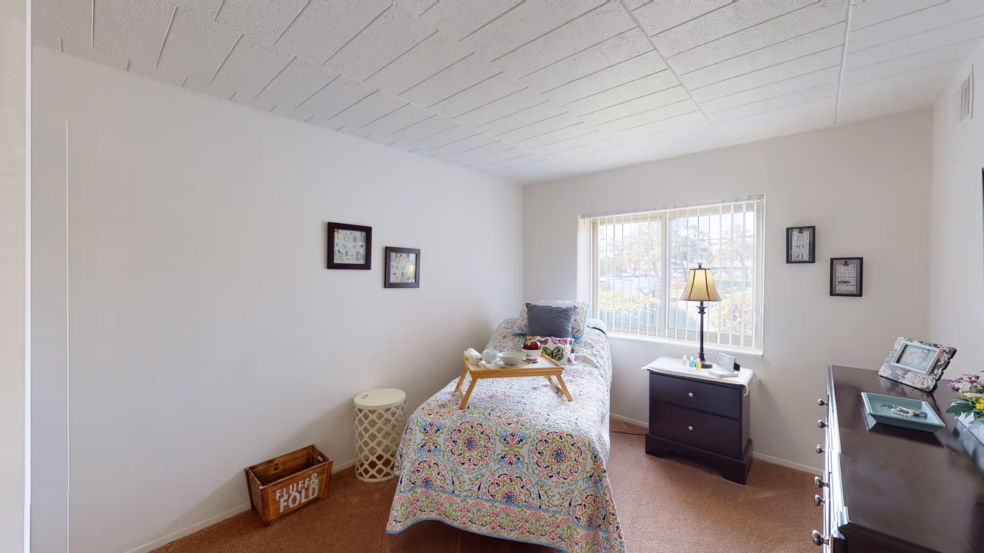 A small, bright bedroom with a single bed covered in a colorful patterned bedspread. A wooden tray with a teapot and cup is placed on the bed. Next to the bed is a dark wooden nightstand with a lamp and some small items. There is a window with vertical blinds letting in natural light. On the opposite side of the room is a dark wooden dresser with framed photos and decorative items. The walls are white with a few small framed pictures hanging.