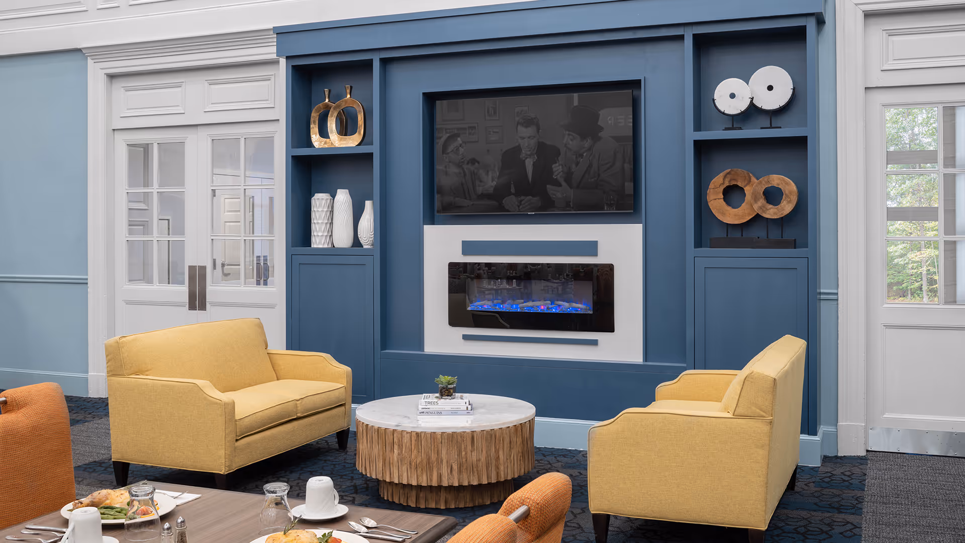 A cozy living room area with two yellow armchairs and a matching loveseat arranged around a round wooden coffee table with a marble top. The background features a built-in blue shelving unit with decorative items, a wall-mounted flat-screen TV showing a black-and-white movie, and an electric fireplace below it. The room has light blue walls and white double doors with glass panes on either side of the shelving unit.