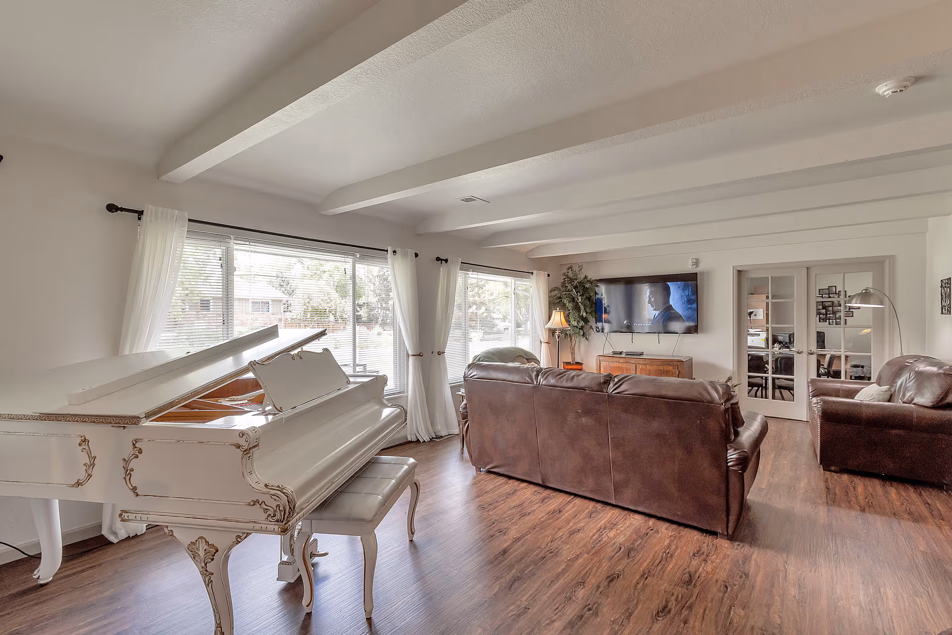 A bright and spacious living room with a white grand piano on the left, large windows with white curtains letting in natural light, a brown leather sofa and armchair facing a wall-mounted flat-screen TV, wooden flooring, and a floor lamp beside the sofa. There are glass double doors in the background leading to another room.