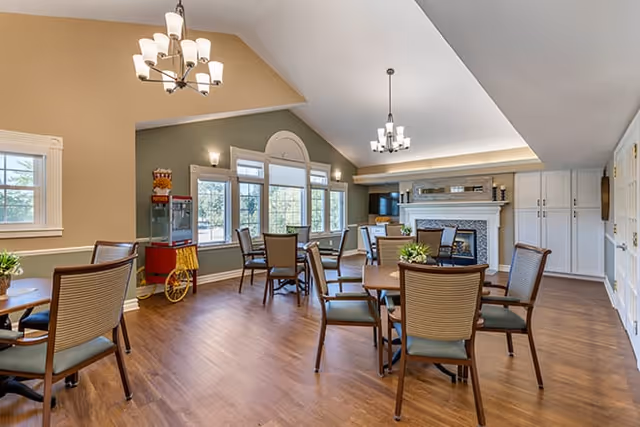A spacious dining room with several round wooden tables and chairs arranged neatly. The room features large windows allowing natural light to fill the space, a popcorn machine in the corner, and a fireplace with a mantel on the far wall. The ceiling has two modern chandeliers, and the floor is covered with wood-style flooring.
