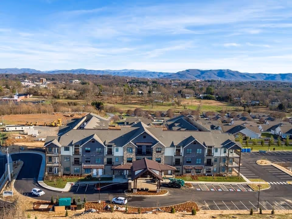 Aerial view of a large senior living facility building with multiple peaked roofs, surrounded by parking lots and landscaped areas. The building is situated in a semi-rural area with fields, trees, and mountains visible in the background under a partly cloudy sky.