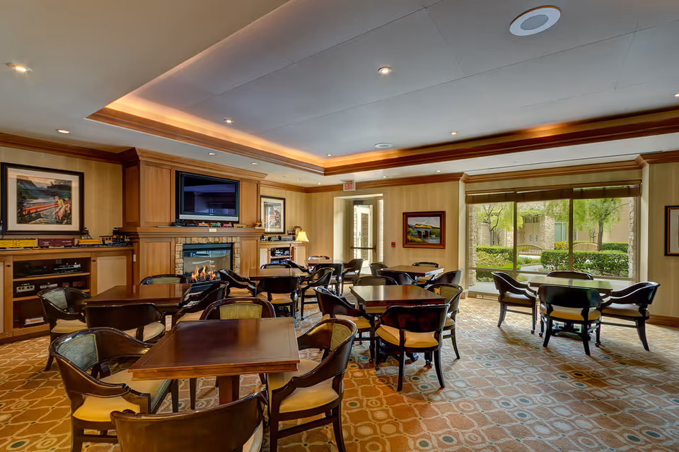 A cozy common area with multiple wooden tables and chairs arranged on a patterned carpet. The room features a fireplace with a TV mounted above it, framed artwork on the walls, and large windows letting in natural light with a view of greenery outside. The ceiling has recessed lighting and a decorative tray design with warm accent lighting.