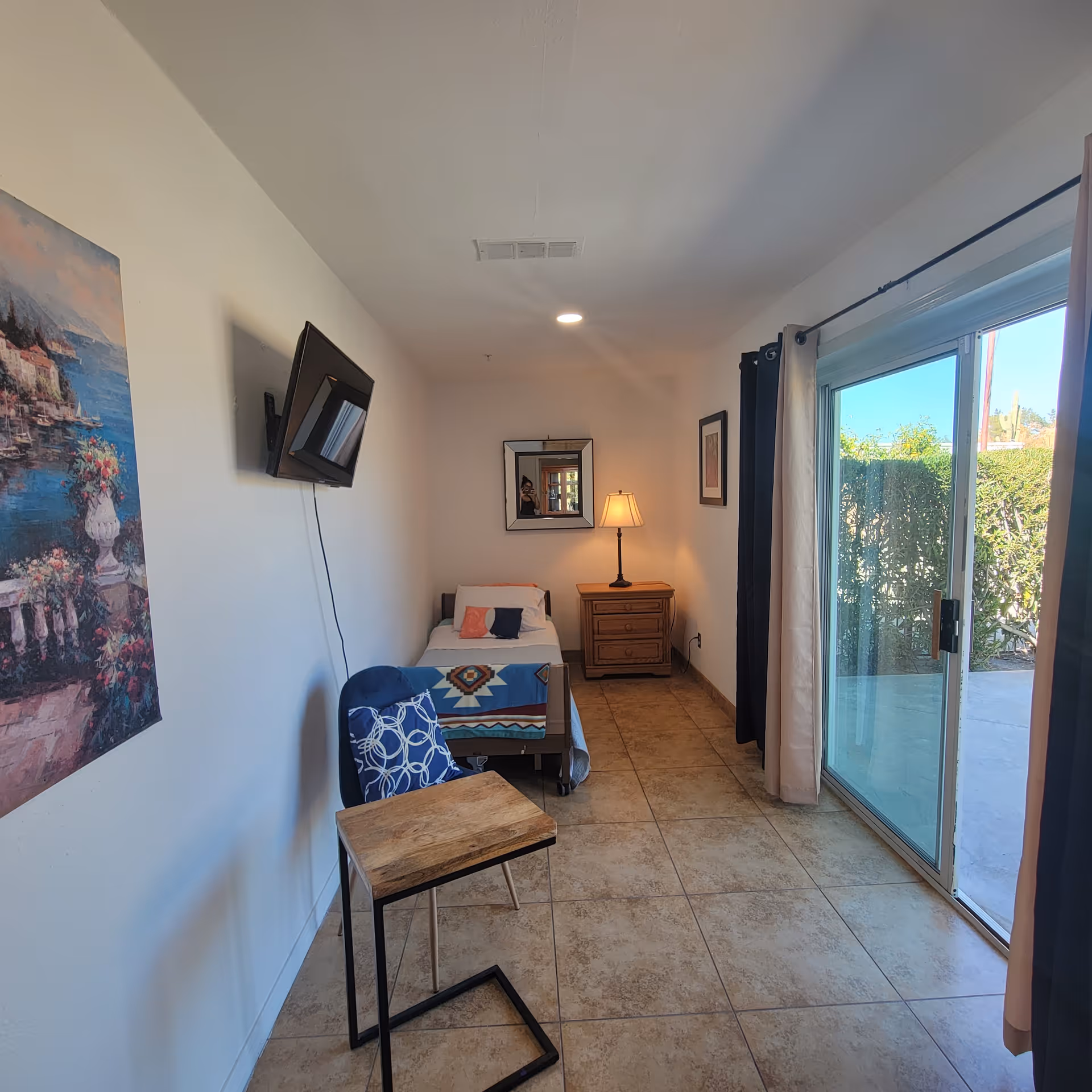A small, tidy bedroom with a single bed covered with a colorful blanket, a wooden nightstand with a lamp, a wall-mounted TV, a blue chair with a patterned pillow, a small wooden table, and a large sliding glass door leading outside. The room has tiled floors and light-colored walls with a painting and a framed picture.