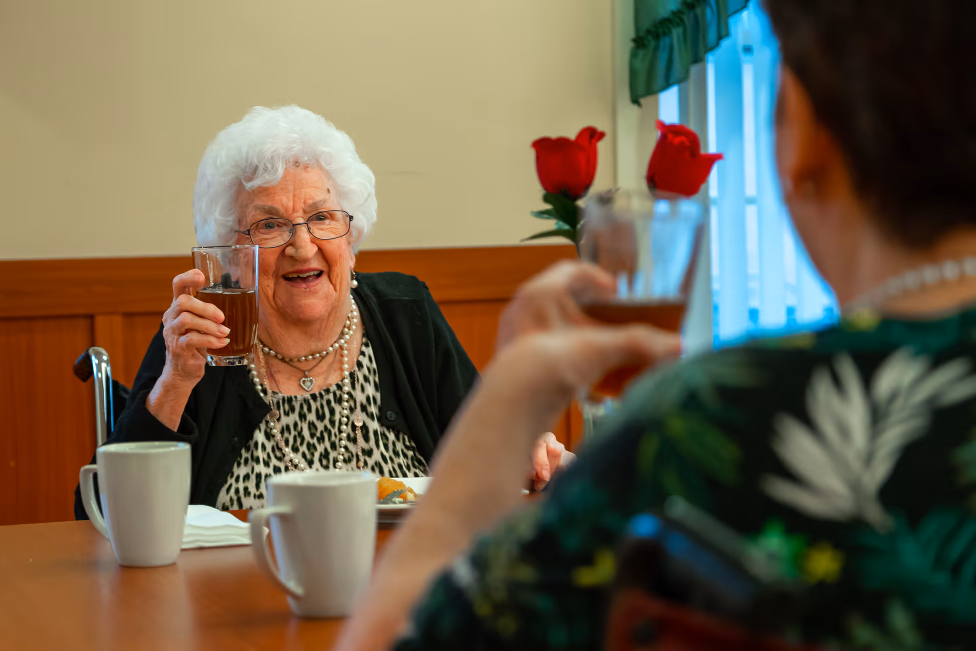 An elderly woman with white hair and glasses is sitting at a dining table, smiling and holding up a glass of a dark beverage in a toast. Another person, partially visible from behind, is also raising a glass. The table has two white mugs and a plate with food. In the background, there are two red roses in a vase near a window with green curtains.