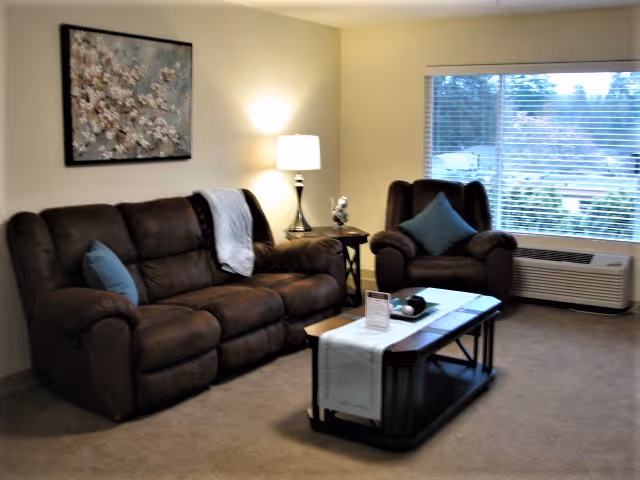 A cozy living room with a brown reclining sofa and matching armchair, both adorned with blue accent pillows. A wooden coffee table with a white runner and decorative items sits in front of the sofa. A side table with a lamp and a small sculpture is placed between the sofa and armchair. A large window with blinds lets in natural light, and a framed floral artwork hangs on the wall above the sofa.