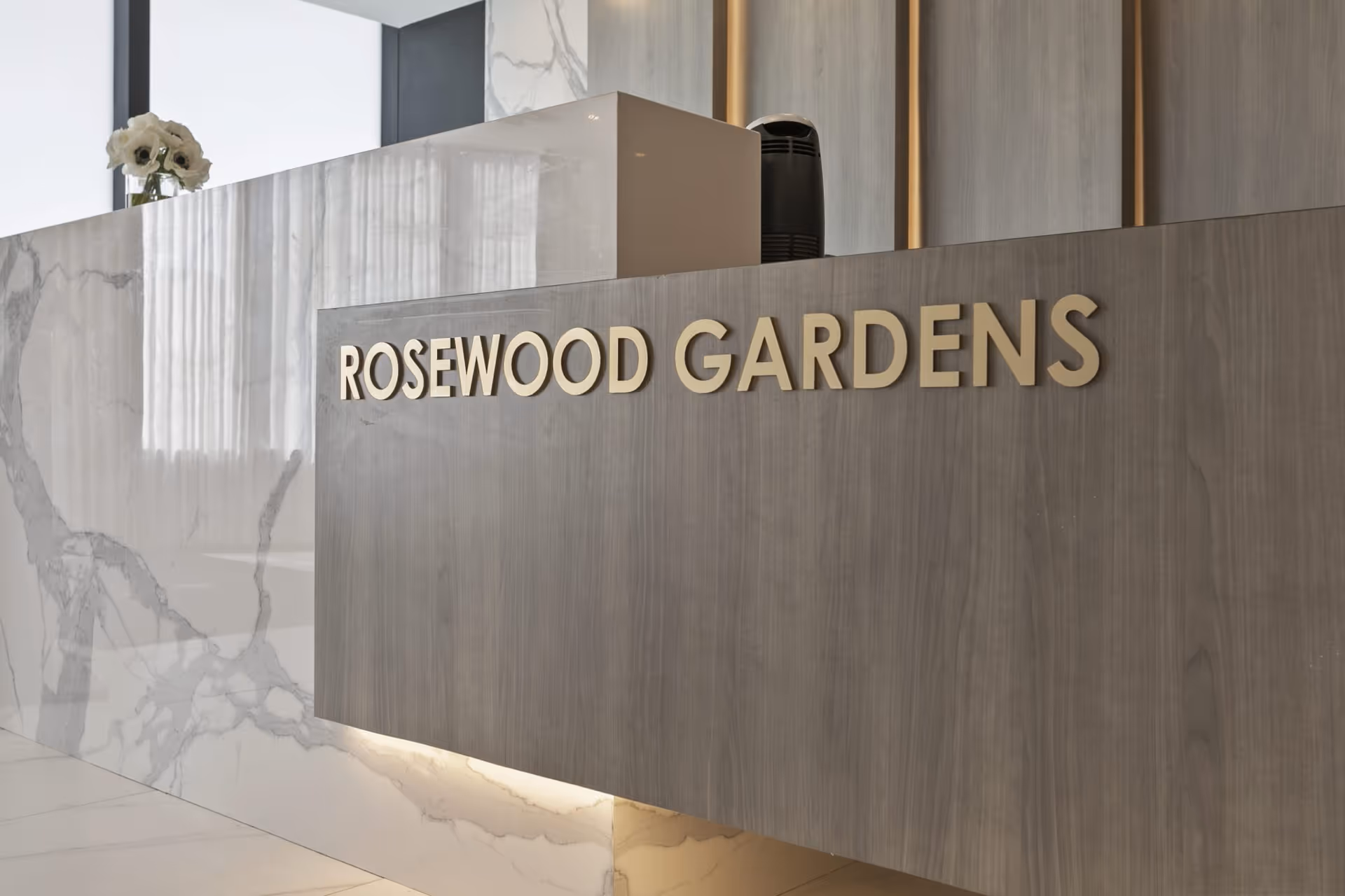 Reception desk area with a modern design featuring a marble countertop and a wooden panel with the text 'ROSEWOOD GARDENS' in gold letters. A small vase with white flowers is placed on the counter.