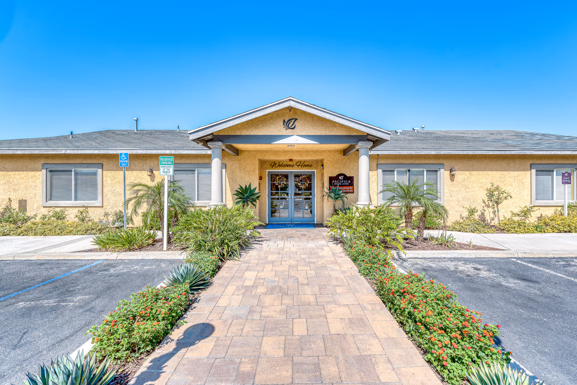 Front exterior view of a single-story senior living facility building with a paved walkway leading to double glass doors under a small portico. The building is beige with a gray roof, surrounded by landscaped plants and bushes. There are signs indicating reserved parking and handicap parking near the entrance. The sky is clear and blue.