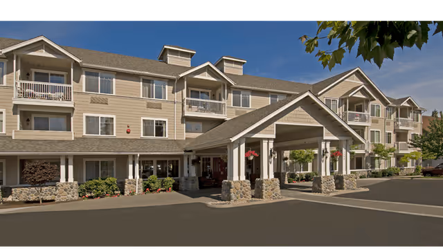 Exterior front view of a three-story senior living building with a covered porte-cochère entrance, balconies, stone columns, and landscaping.