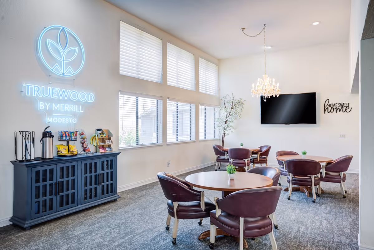 A bright and airy common area with several round tables and purple cushioned chairs on wheels. Large windows with white blinds let in natural light. A chandelier hangs from the ceiling, and a flat-screen TV is mounted on the wall next to a decorative sign that reads 'Home Sweet Home.' A dark blue cabinet holds coffee supplies and snacks beneath a neon sign that says 'Truewood by Merrill Modesto.'