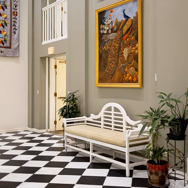 Decorative lobby with a white bench, potted plants, a large peacock painting, and a black-and-white checkered floor.