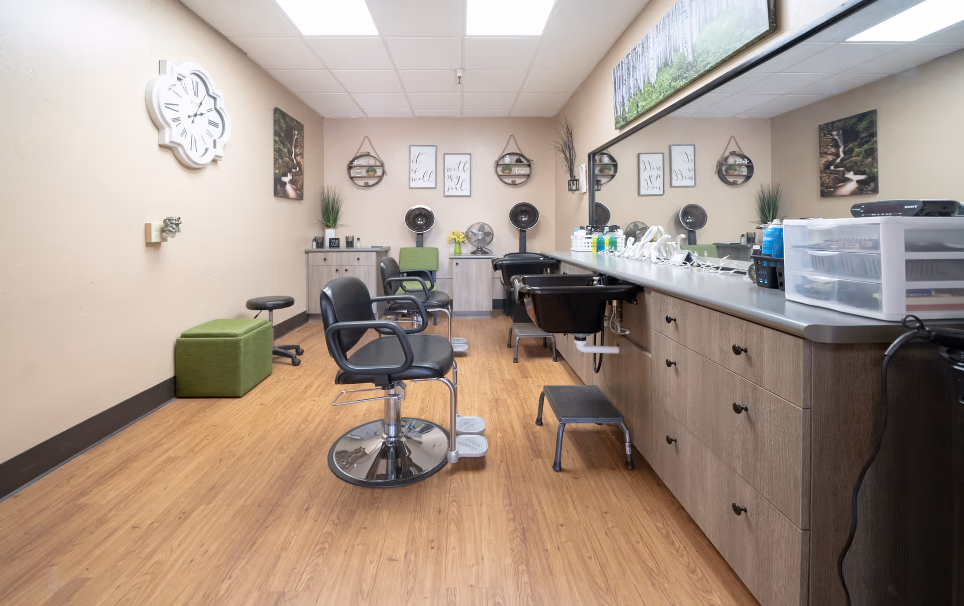 Interior view of a hair salon area in a care center with wooden flooring, two black salon chairs, a green chair, a green cushioned bench, and a counter with sinks and hair care products. The walls are decorated with framed pictures, plants, and a large clock.
