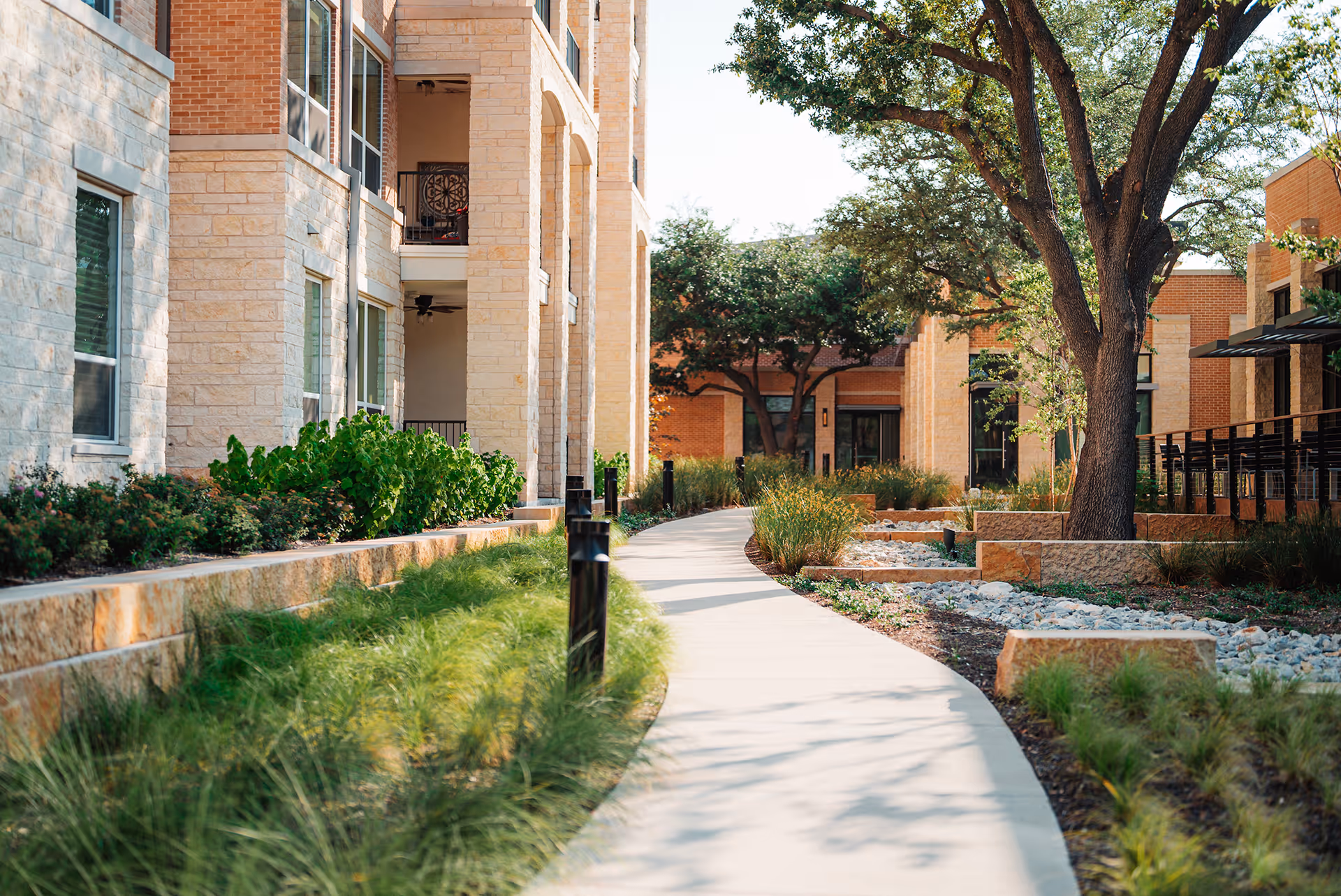 A winding concrete pathway surrounded by landscaped greenery and trees, flanked by multi-story buildings with stone and brick exteriors under a clear sky.