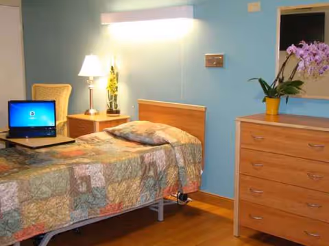 Bedroom in a senior living facility with a bed, dresser, laptop, and orchid plant.