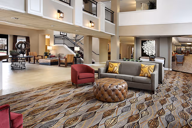 Spacious senior living facility common area with modern furniture including a gray sofa with yellow pillows, red armchairs, and a large tufted leather ottoman on a patterned carpet. The area features high ceilings, wall sconces, large windows, and a staircase leading to an upper level. In the background, there is additional seating and a decorative sculpture on a table.