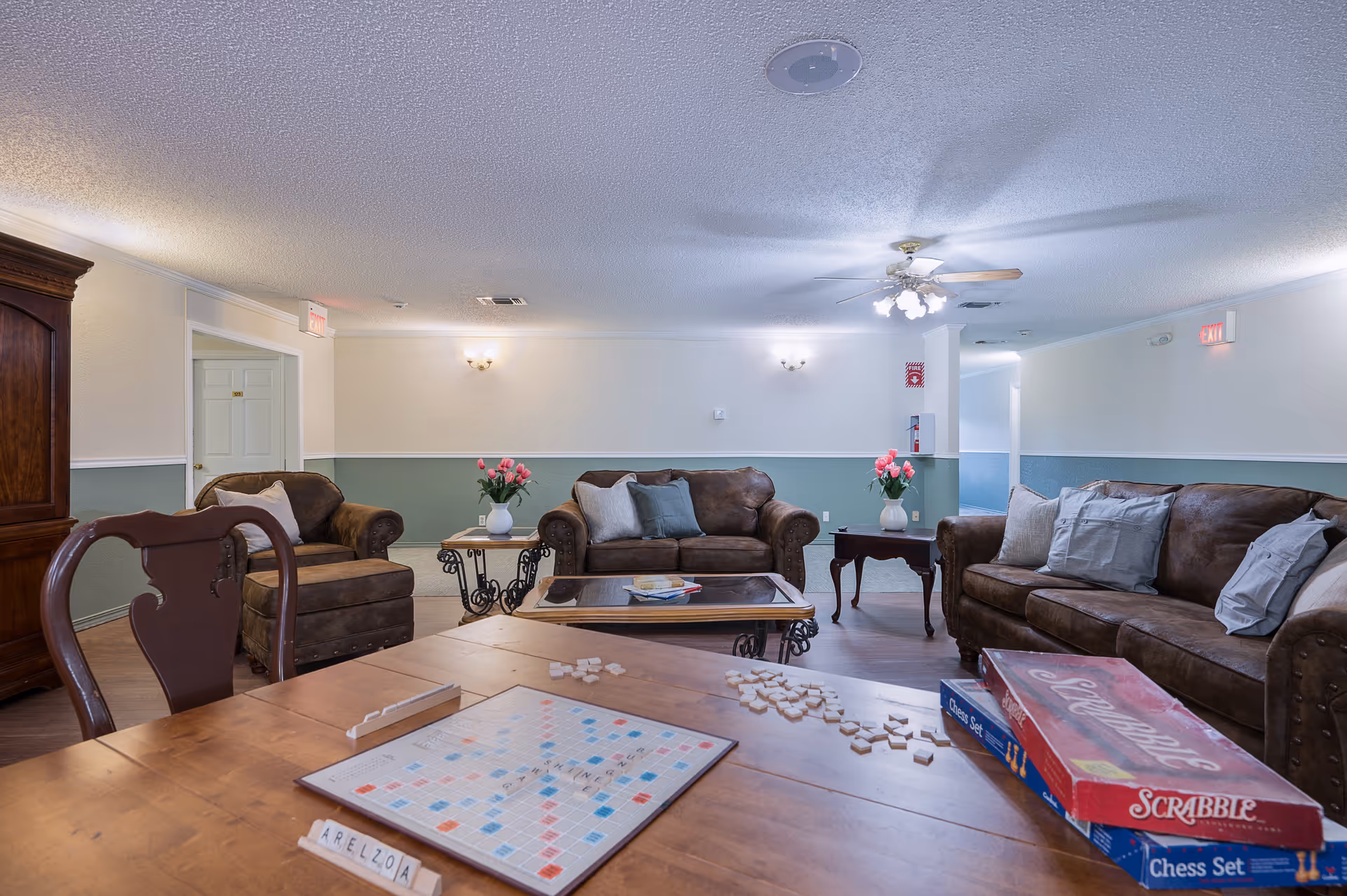 A cozy common area in a senior living facility with brown leather sofas, a wooden coffee table, and side tables with vases of pink flowers. A ceiling fan with lights is mounted on the ceiling. In the foreground, a wooden table holds a Scrabble board game and a chess set box, with Scrabble tiles scattered on the table. The walls are painted white and light green with wall sconces providing additional lighting.
