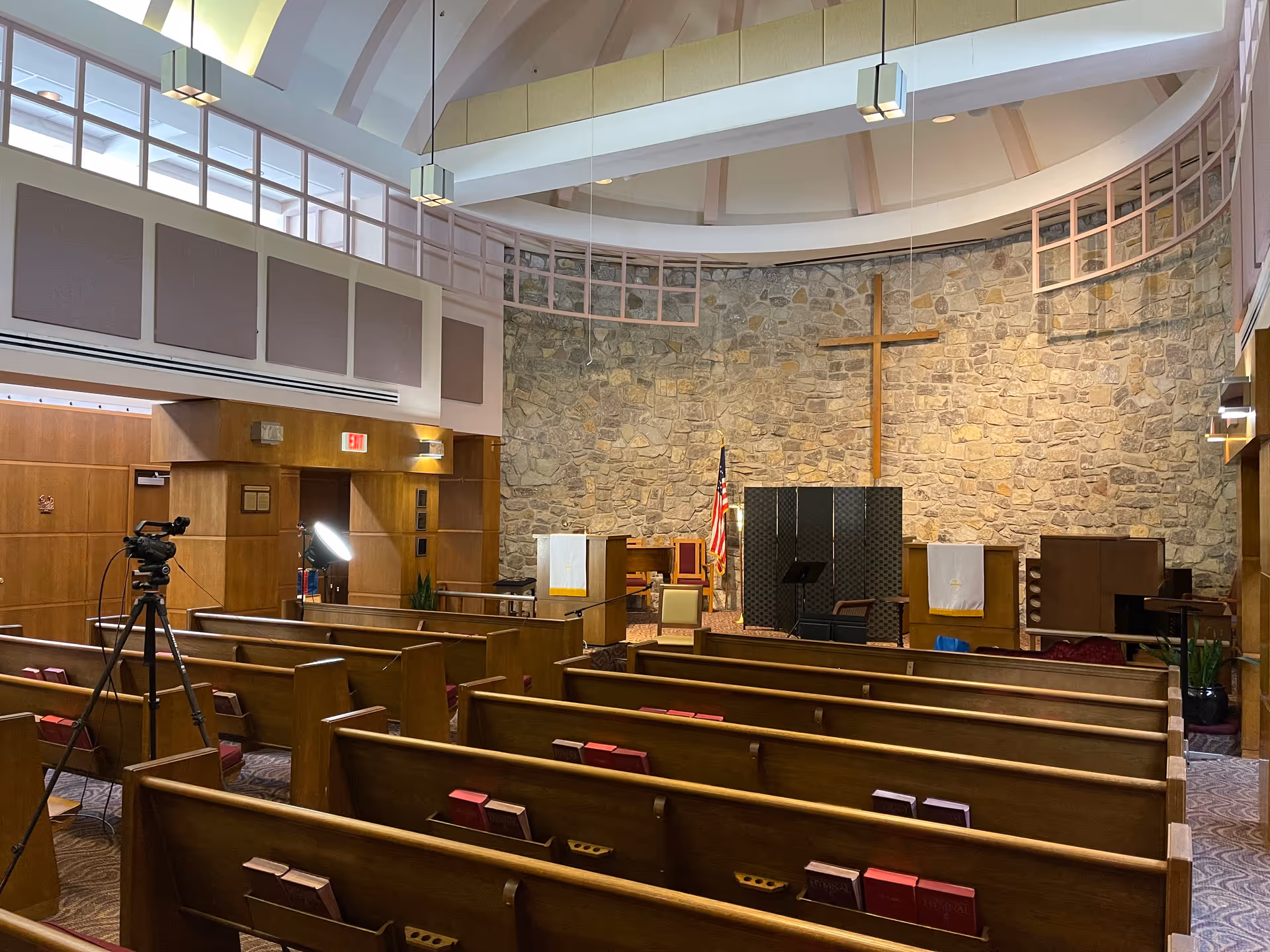 Interior view of a chapel with wooden pews, a stone wall with a large wooden cross, an American flag, and a lectern. The ceiling has a circular design with hanging lights. A camera on a tripod and a lighting setup are visible on the left side.