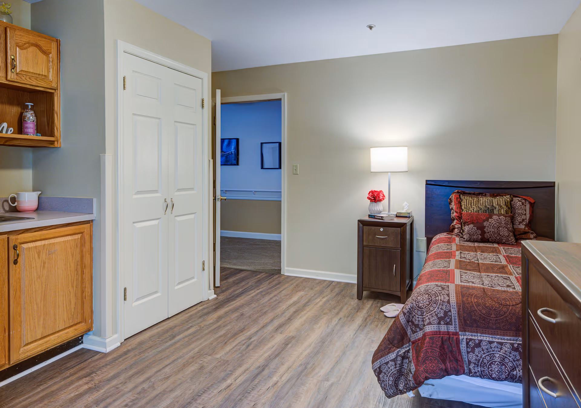 A small bedroom with a single bed covered in a patterned red and brown quilt. Next to the bed is a dark wooden nightstand with a lamp, a small vase with red flowers, and some books. The room has light-colored walls and wood flooring. To the left, there is a kitchen area with wooden cabinets and a countertop. A white double-door closet and an open door leading to another room are also visible.