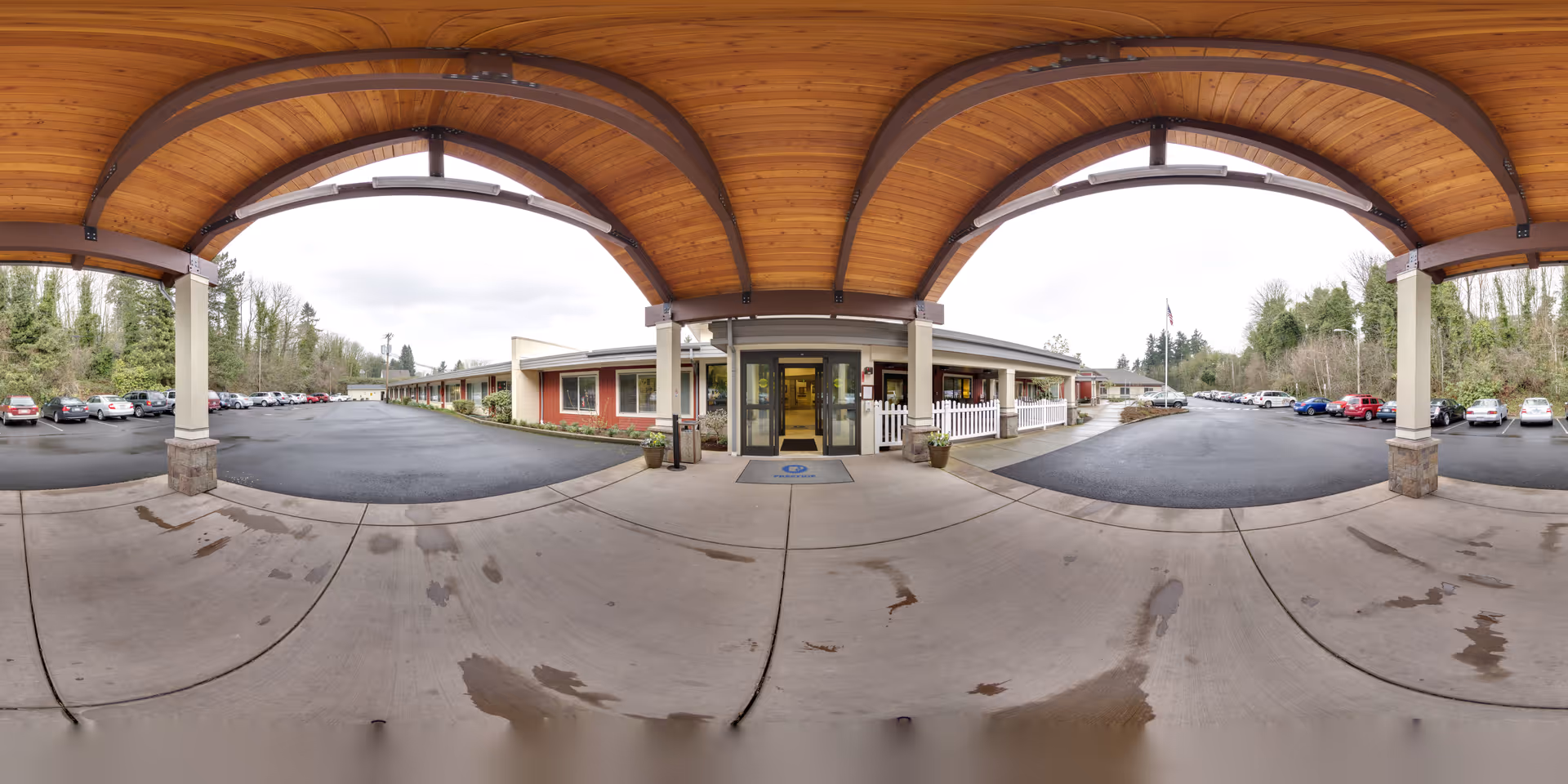 Covered entrance area with wooden ceiling and concrete floor leading to the main entrance of a single-story building. The building has red and beige exterior walls with windows and a white picket fence section. There are parking lots with multiple cars on both sides of the entrance, surrounded by trees and greenery under an overcast sky.