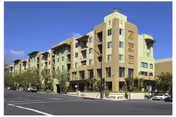 Exterior view of a multi-story senior living facility building with a mix of beige and green colors, featuring balconies and windows, situated on a street corner with trees and parked cars.