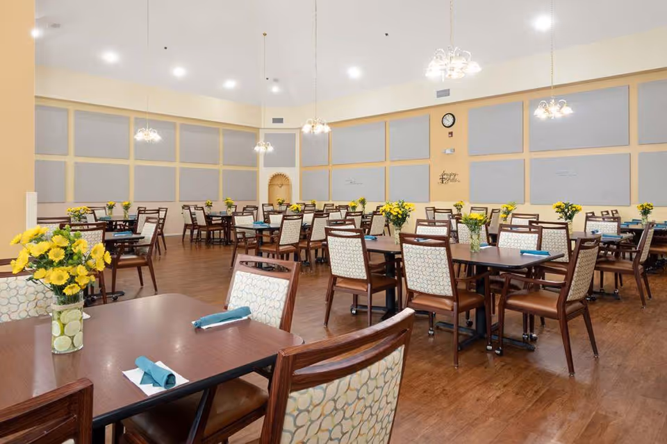 Large communal dining room with wooden tables and upholstered chairs arranged with yellow floral centerpieces and hanging chandeliers.