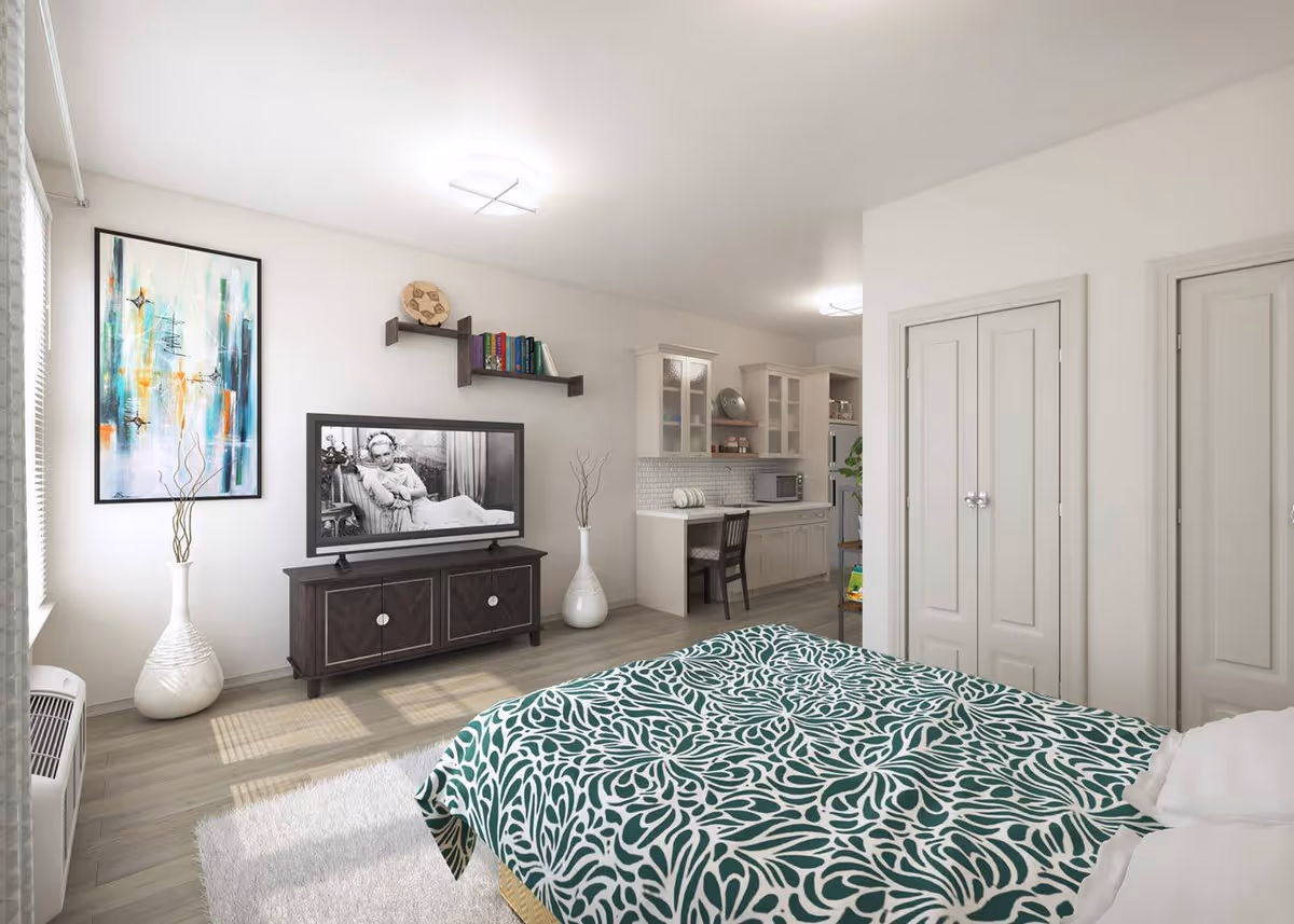Bright studio-style bedroom with a patterned bedspread, TV on a console, decorative vases and wall art, and a small kitchenette in the background.