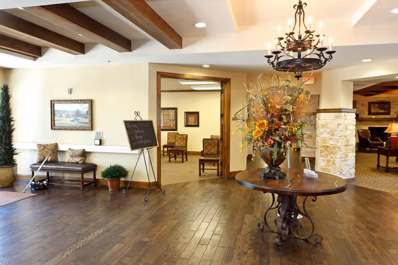 A spacious and warmly decorated interior common area with wooden floors, a round wooden table with a large floral arrangement, a chandelier, a bench with cushions, framed artwork on the walls, and an open doorway leading to a room with chairs and additional framed pictures.