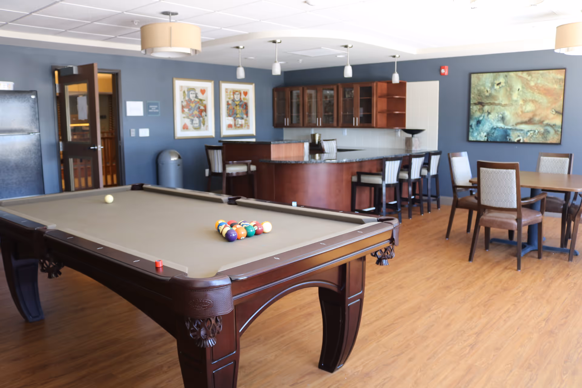 A recreational room with a pool table in the foreground, a bar area with stools in the background, and a dining table with chairs to the right. The walls are painted blue and decorated with framed artwork. The floor has a wood finish.