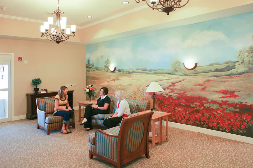 A cozy sitting area in a senior living facility with three women engaged in conversation. The room features striped upholstered chairs and a sofa, a side table with a lamp, and a large mural on the wall depicting a scenic field with trees and a blue sky. A chandelier hangs from the ceiling, and there is a small cabinet with a plant and books near the door.