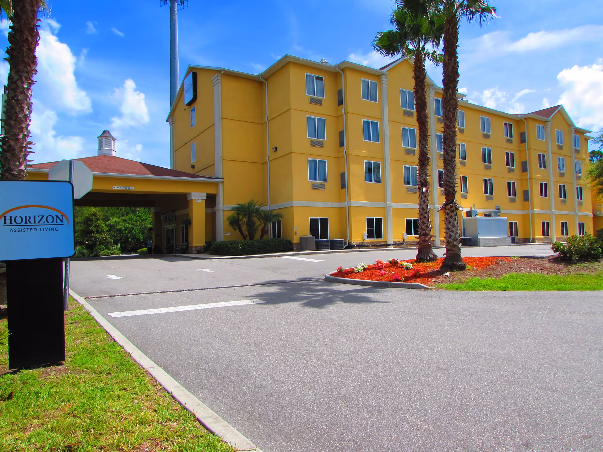 Exterior view of a four-story yellow assisted living facility building with multiple windows, palm trees, and a small landscaped area with flowers in front. A sign near the entrance reads 'Horizon Assisted Living'. The sky is blue with some clouds.
