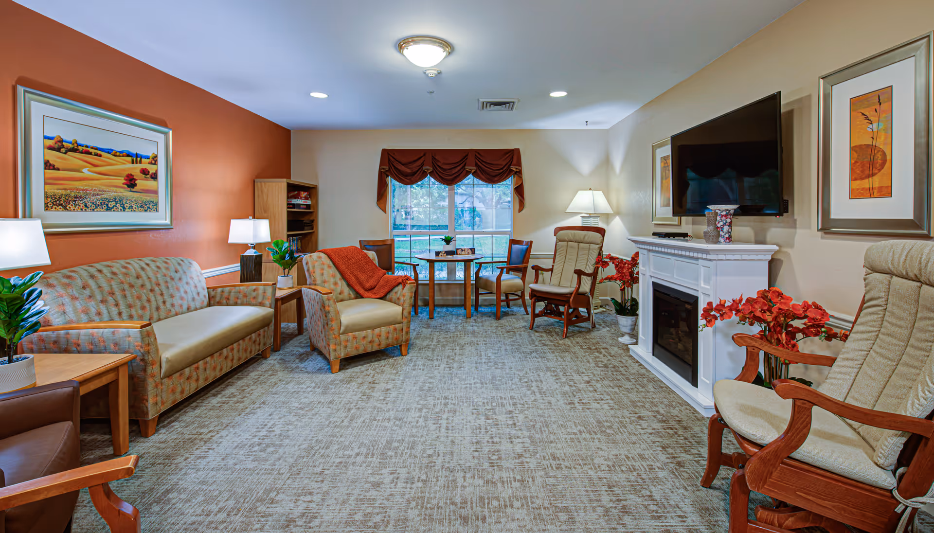 A cozy living room with patterned sofas and armchairs arranged around a white electric fireplace with a mounted flat-screen TV above it. The room has a large window with a brown valance, a small round table with two chairs in front of the window, and various decorative plants and framed artwork on the walls. The carpet is light-colored with a subtle pattern, and the walls are painted in warm tones including a burnt orange accent wall.