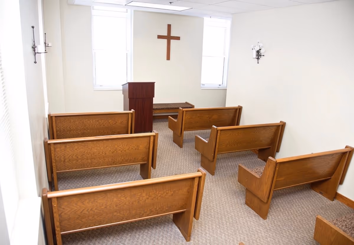 Small chapel room with wooden pews arranged in rows facing a wooden podium. A wooden cross is mounted on the wall between two windows. The room has beige walls and carpeted floor, with wall-mounted candle holders on either side.