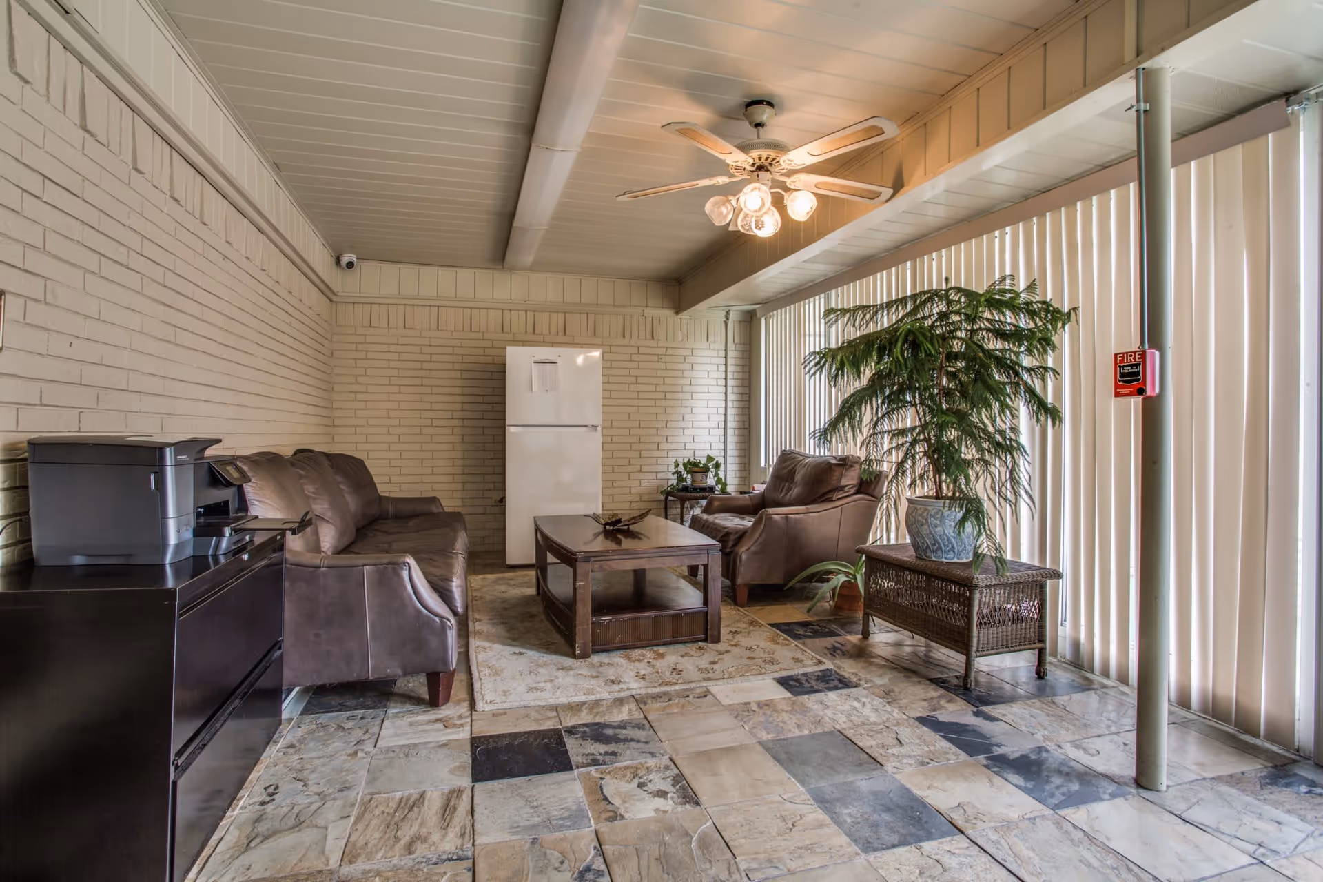 A cozy indoor seating area with a brown leather sofa and armchair, a wooden coffee table on a patterned rug, a large potted plant on a wicker table, a white refrigerator against a brick wall, a black printer on a black cabinet, and vertical blinds covering the windows. The ceiling has a white fan with lights.