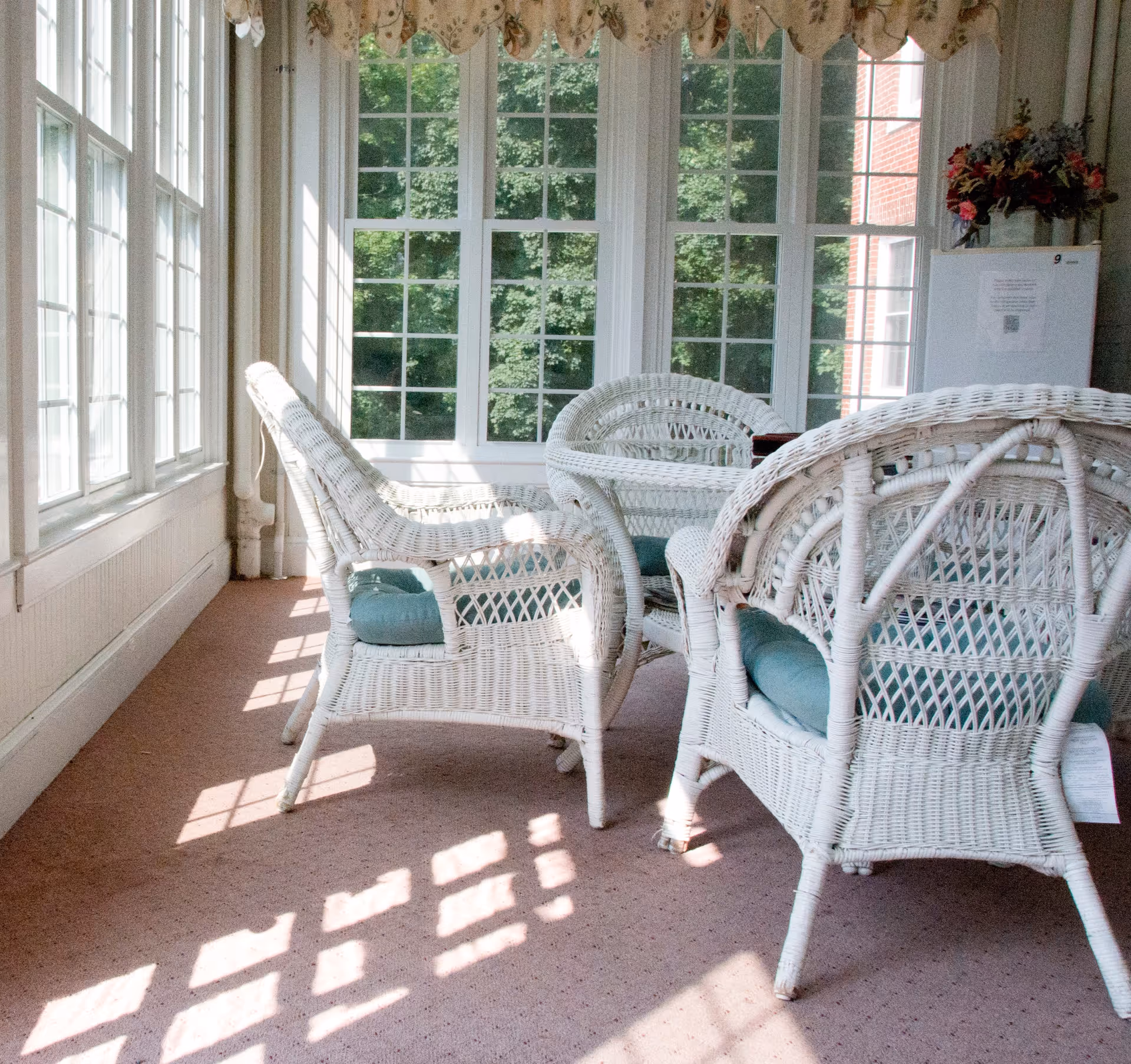 A sunlit room with large windows showing green trees outside. Inside, there are white wicker chairs with blue cushions arranged around a small table. A white cabinet with a flower arrangement on top is visible in the corner.