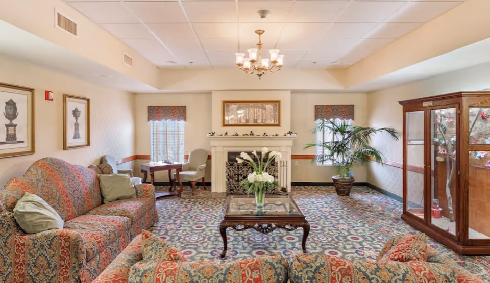 A cozy living room area in a senior living facility featuring patterned upholstered sofas with cushions, a wooden coffee table with a vase of white flowers, a fireplace with decorative items on the mantel, framed artwork on the walls, two windows with patterned valances, a potted plant, and a wooden display cabinet with glass doors containing decorative items.