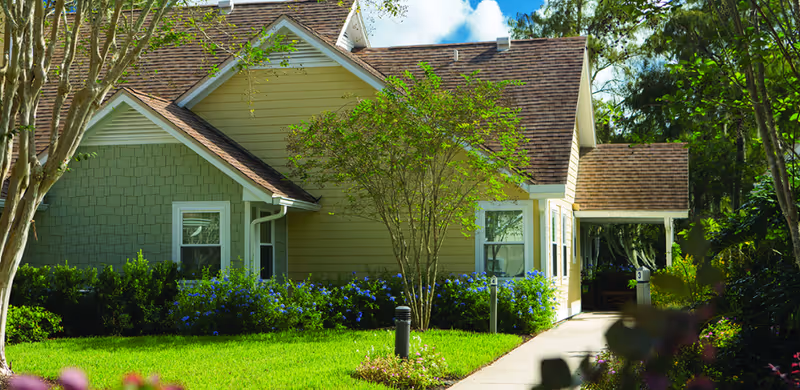 Front exterior of a single-story residential building with a gabled roof, walkway, lawn, and landscaped shrubs and trees.