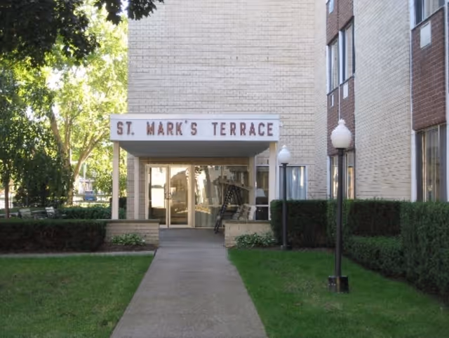 Entrance of St. Mark's Terrace building showing a covered walkway with a sign, glass doors, and surrounding lawn and shrubs.