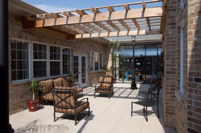 Sunlit brick courtyard patio with a wooden pergola, cushioned lounge chairs, potted plants, and glass doors.