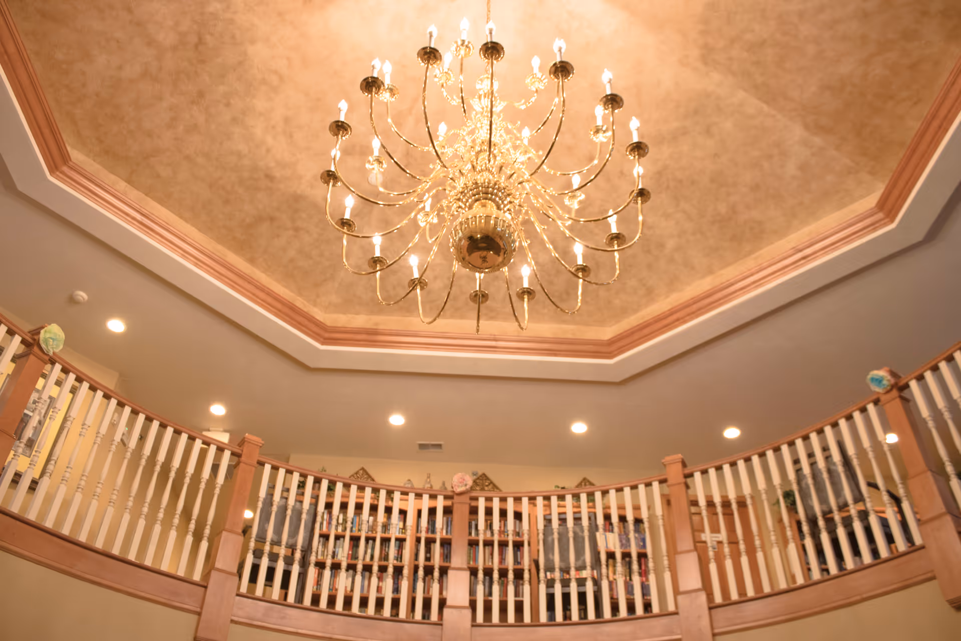View of an elegant chandelier hanging from a decorative recessed ceiling above a curved balcony railing with bookshelves and decorative items in the background.