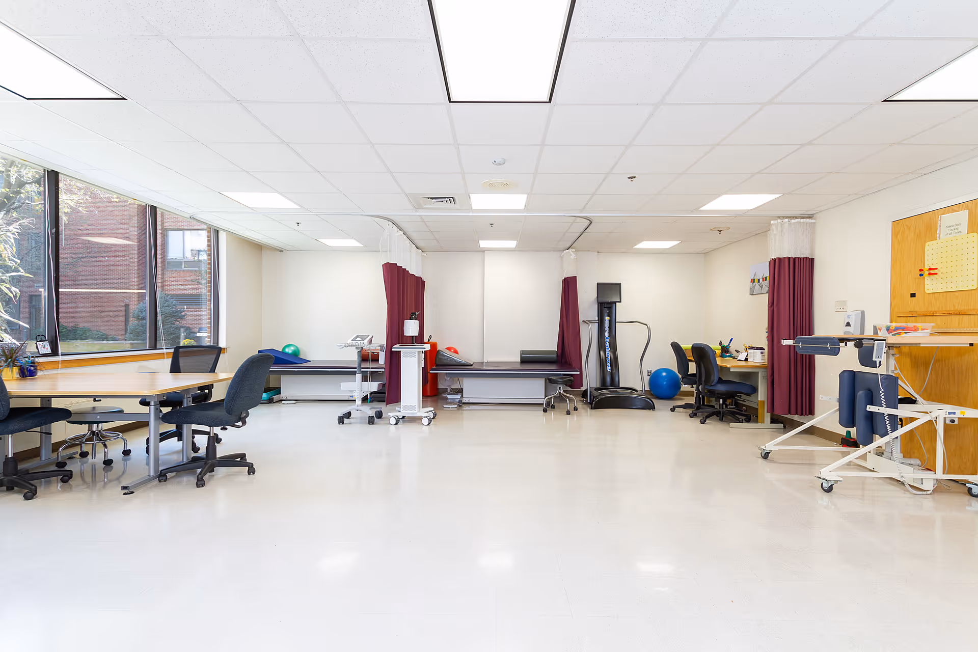 A bright and spacious rehabilitation room with large windows on the left side letting in natural light. The room contains several office chairs, tables, medical examination beds with privacy curtains, exercise equipment including a vibration plate and therapy balls, and a corkboard on the right wall with notes pinned to it.