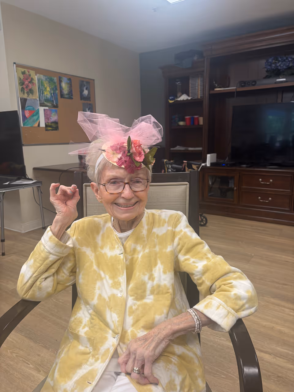 An elderly woman wearing glasses and a yellow tie-dye jacket sits in a chair indoors. She is smiling and wearing a pink floral headpiece with tulle. Behind her is a wooden entertainment center with a TV, shelves, and various items, as well as a bulletin board with artwork on the wall.
