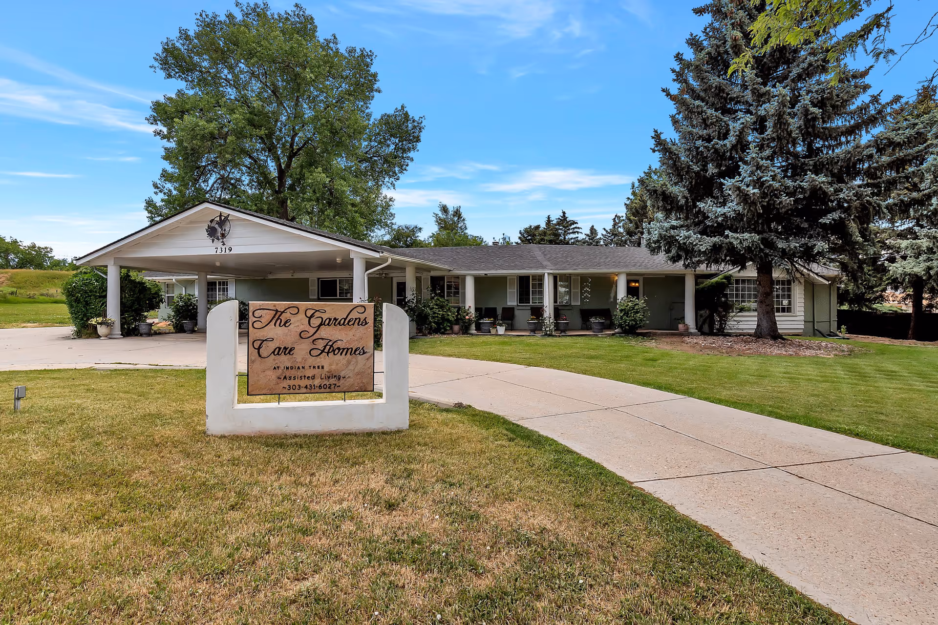 Exterior view of a single-story assisted living facility named The Gardens Care Homes at Indian Tree, featuring a driveway, green lawn, large trees, and a sign with the facility's name and contact number.