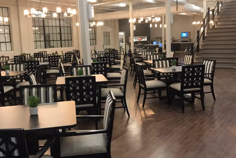 Spacious dining room with rows of dark wood tables and cushioned chairs, overhead chandeliers, and a staircase in the background.