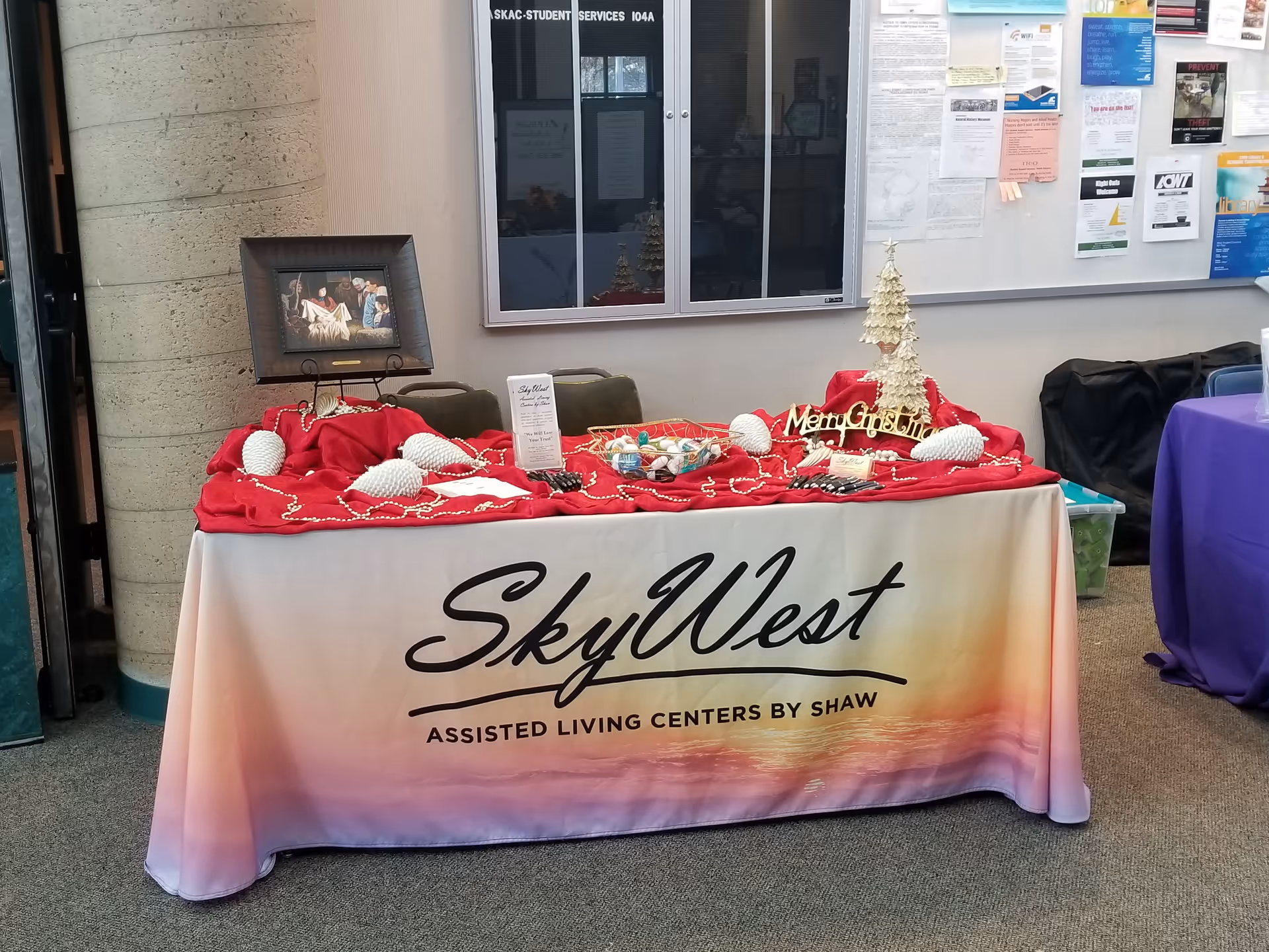 A promotional table for SkyWest Assisted Living Centers by Shaw, covered with a red cloth and decorated with white pinecone ornaments, a small white Christmas tree, and a 'Merry Christmas' sign. Behind the table is a bulletin board with various notices and flyers.